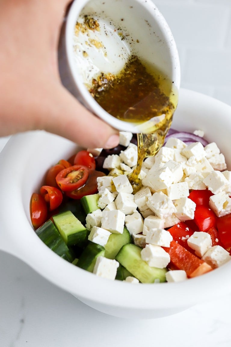 shot of homemade greek dressing being poured into white bowl with english cucumbers, grape tomatoes and feta cheese