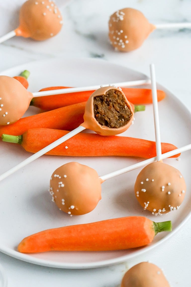 4 carrots and 4 cake pops laying down on a white plate with one cake pop in half