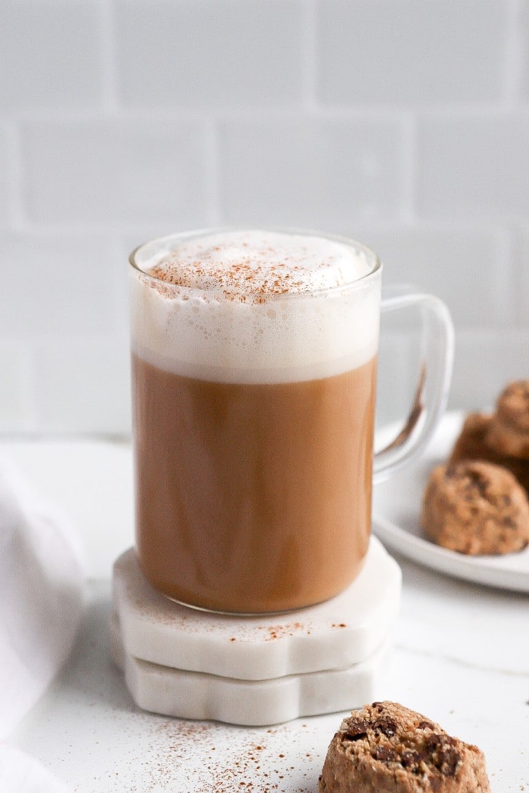 a skinny latte in a clear mug with white foam and cinnamon with treats behind it