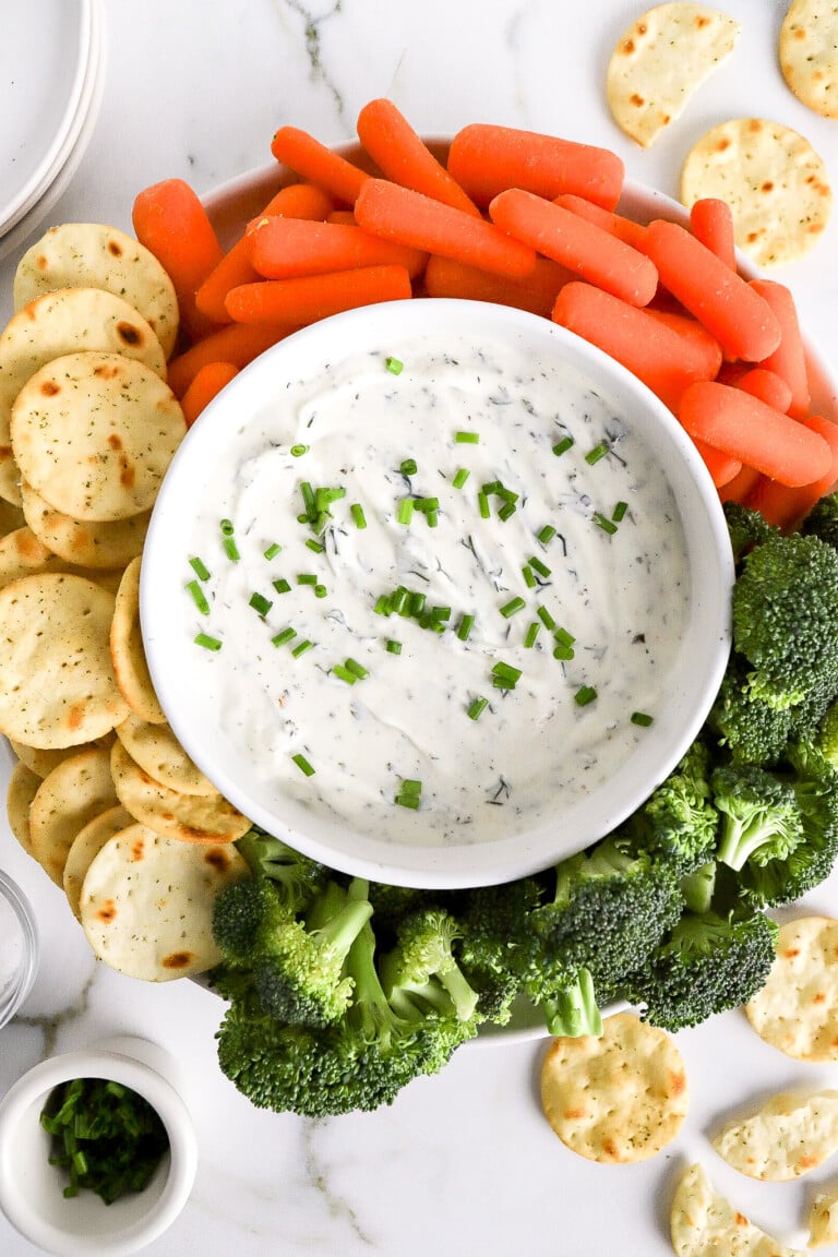 Top down view of homemade greek yogurt ranch dip with carrots, broccoli and crackers on white plate.