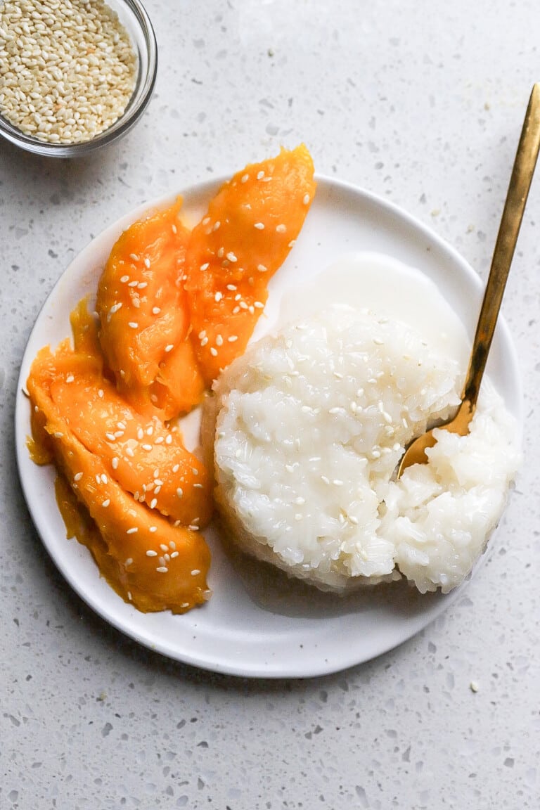 Top down view of thai mango sticky rice with gold spoon and a side of white sesame seeds.