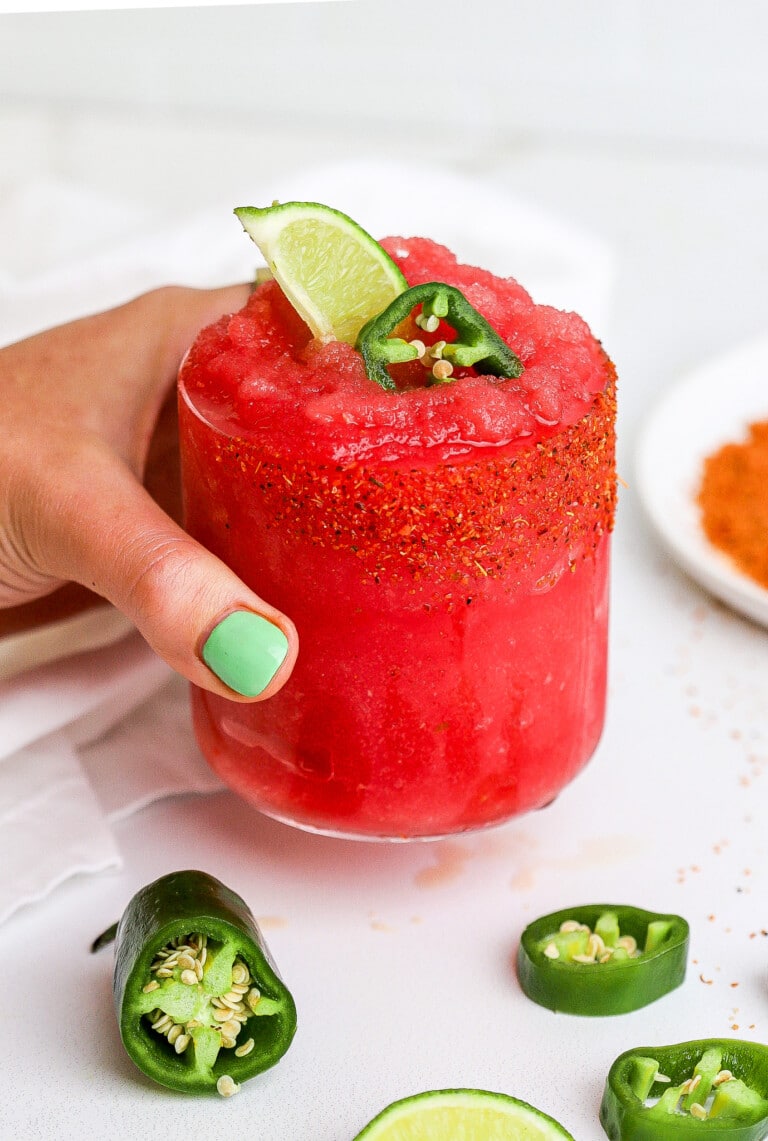 Side angle of a hand holding a frozen margarita with a jalapeno slice and lime wedge sticking out of the glass.