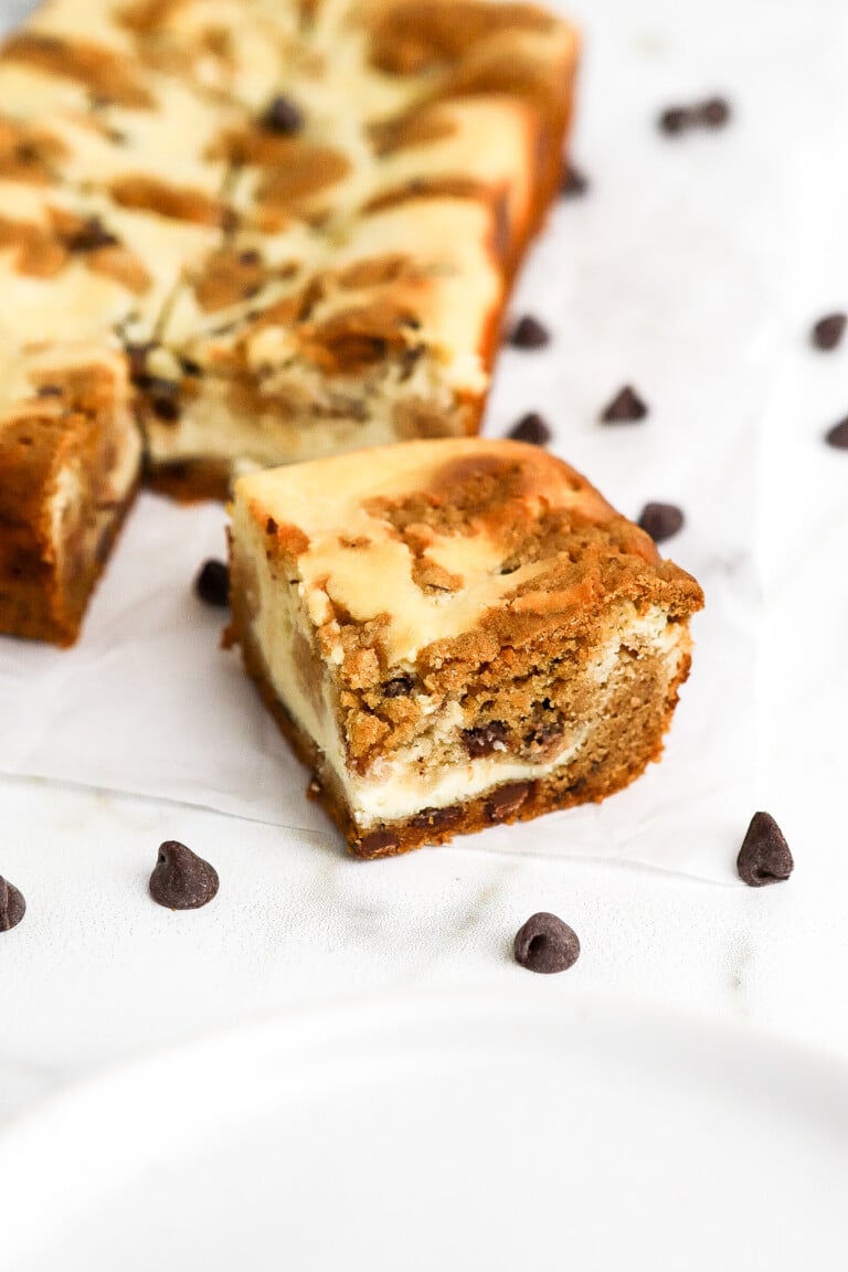 Side angle of easy cookie dough cheesecake bars with chocolate chips scattered around with small white plates in foreground.