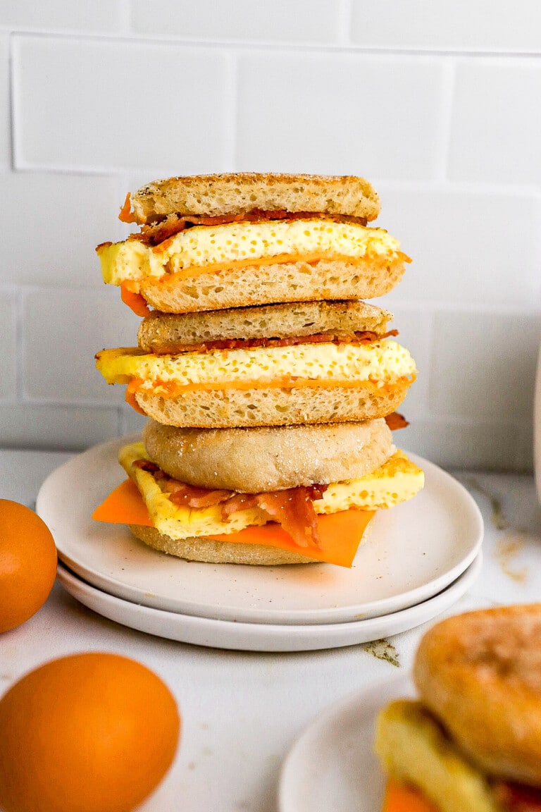 3 freezer bacon breakfast sandwiches stacked on top of one another on white plate with eggs in foreground.