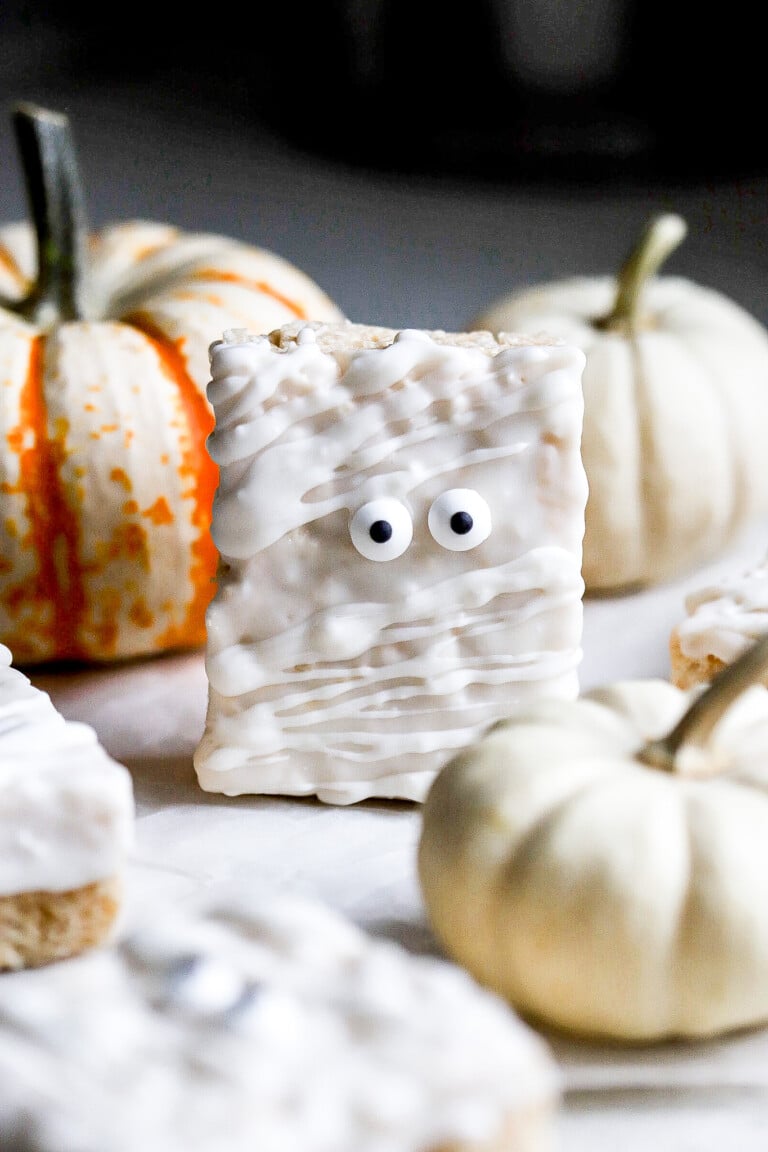 Close up view of mummy Rice Krispie treats with candy eyeballs on parchment paper with pumpkins in background.