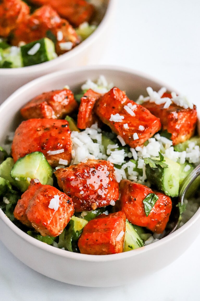 Side shot of cubes of hot honey salmon in white bowl with white rice and avocado and cucumbers.