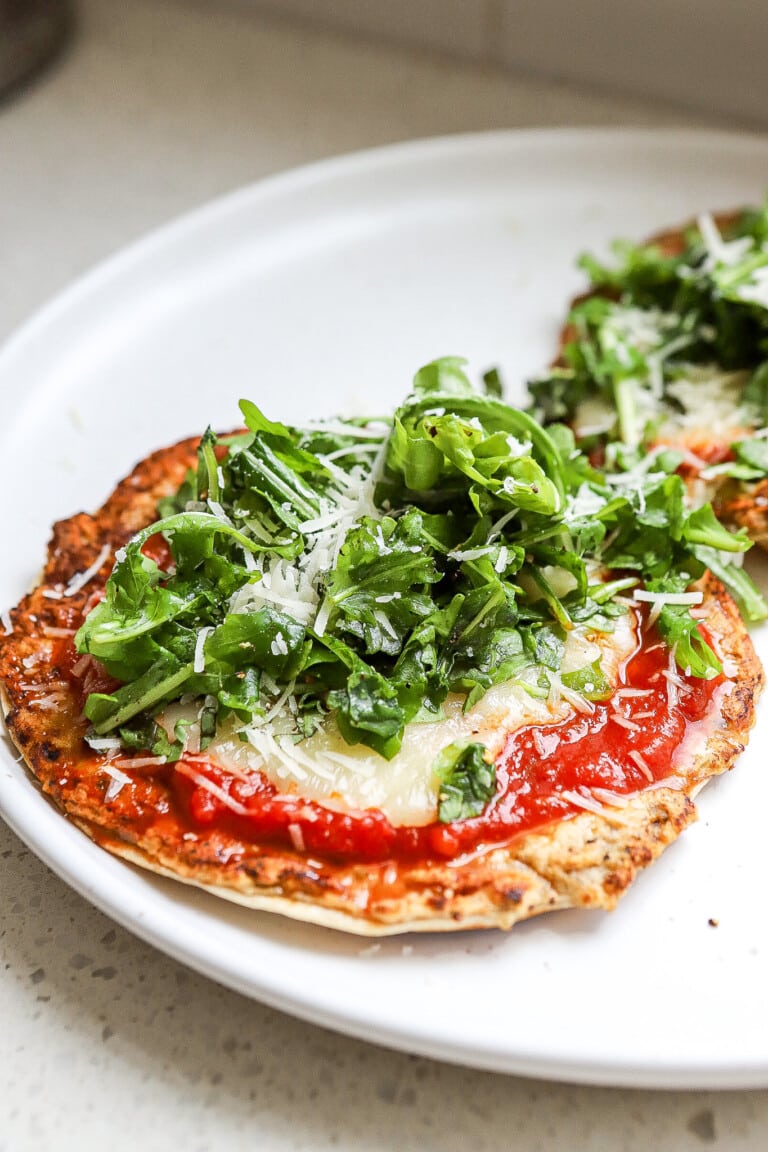 Side angle of chicken parm smash tacos on a small white plate with fresh arugula on top.