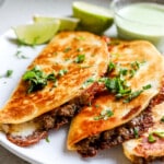 Side angle of crispy beef tacos with cilantro lime crema in background.